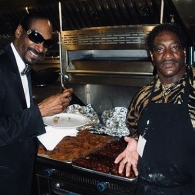 Snoop is eating chicken while Reo is on an apron.